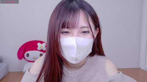 Media: Video of an Asian woman with long, straight brown hair, wearing a white face mask and beige off-shoulder sweater, in a room with a pink Hello Kitty plush toy and white walls.