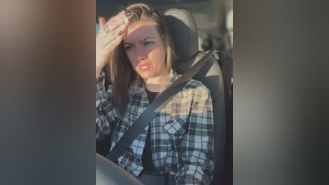 Media: Video of a Caucasian woman with long brown hair, wearing a plaid shirt, seated in a car with a seatbelt, looking tired and holding her head, sunlight filtering through the window.