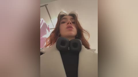 Media: A video of a young woman with long, wavy auburn hair, wearing a white coat and black headphones, looking upward with a neutral expression. The background is blurred, showing a white wall and a ceiling light.