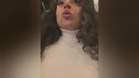 Media: A close-up video of a young woman with curly brown hair and light skin, wearing a ribbed white turtleneck sweater. She has full lips and is blowing a kiss.