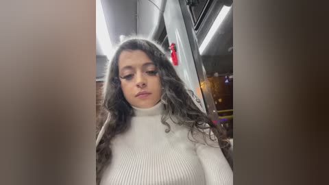 Media: Video of a young woman with long, wavy brown hair, wearing a white ribbed turtleneck sweater, standing inside a subway car. Background shows metal railings, fluorescent lights, and an open door.