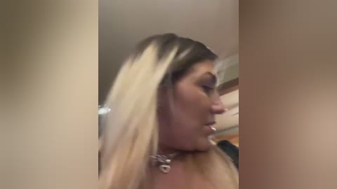 Media: A blurred video of a light-skinned woman with long, blonde hair, wearing a silver necklace, captured from a side profile, indoors with beige walls.