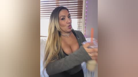 Media: A video of a Latina woman with long, blonde hair, wearing a low-cut, green top, and a choker necklace, sitting on a bed with white sheets, holding a pink drink.