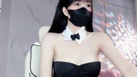 Media: A video of an East Asian woman with long black hair, wearing a black strapless dress, black mask, and white bow tie, seated in a white chair against a marble wall.