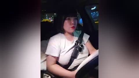 Media: Video of a young woman with black hair and pale skin wearing a white T-shirt, sitting in a car with a black seatbelt, driving at night.