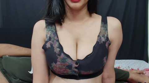 Media: Video of an Asian woman with medium skin tone, wearing a black floral-patterned bra that accentuates her large breasts. Her dark hair is shoulder-length, and she sits on a bed with a dark curtain background.