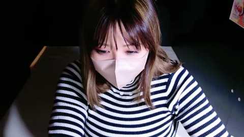 Media: Video of a woman with straight, shoulder-length brown hair, wearing a black-and-white striped sweater, and a white mask, looking down, in a dimly lit room with a dark background.