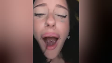 Media: A close-up video of a young woman with fair skin, dark hair, and visible tongue piercing, eyes closed, mouth open, and face slightly flushed, with a blurred background.