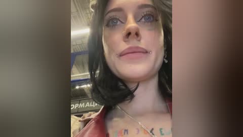 Media: Video of a pale-skinned woman with black hair, blue eyes, and a septum piercing. She wears a red leather jacket and has tattoos on her chest. The background shows an industrial setting with metal beams.