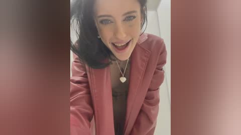 Media: Video of a smiling woman with fair skin, dark hair, and blue eyes, wearing a red blazer over a brown top. She has a heart pendant necklace. The background is blurred, focusing on her cheerful expression.