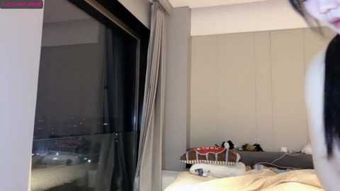 Media: Video of a modern, minimalist bedroom with beige walls, a bed with white linens, and a decorative pillow on the floor. A window with sheer curtains and a partially visible cityscape.