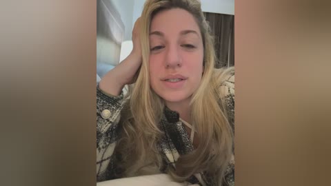 Media: Video of a blonde woman with long hair, wearing a plaid shirt, resting her head on her hand, appearing relaxed, in a dimly lit room.