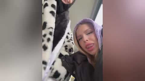 Media: Video of a woman in a cow print outfit and purple hood, sitting in a car, with a blurred car interior visible.