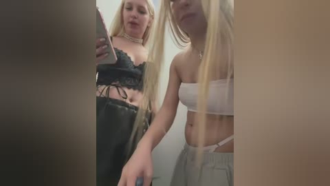 Media: Video of two young women with long blonde hair, one in a black lace bralette and high-waisted shorts, the other in a white bandeau top and loose gray pants. They're indoors, possibly in a bathroom, holding a phone and mirror.