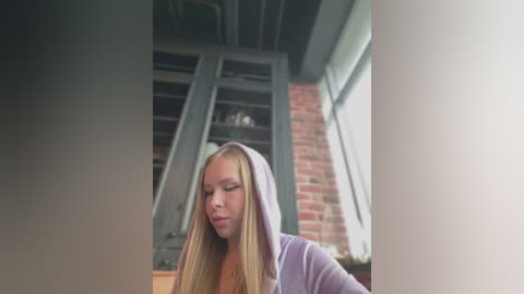 Media: Video of a blonde woman in a lavender hoodie, sitting indoors by a window, with brick wall and bookshelf background.