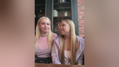 Media: Video of two blonde women, one with long hair, the other with short hair, wearing light-colored shirts, seated in a brick-walled caf\u00e9, smiling at each other.