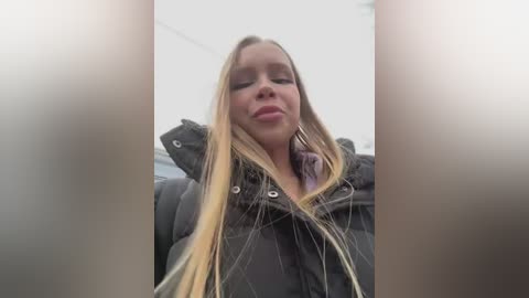 Media: Video of a blonde woman with long hair, wearing a puffy black jacket, giving a slight pout. The background is blurry, featuring an indoor setting.