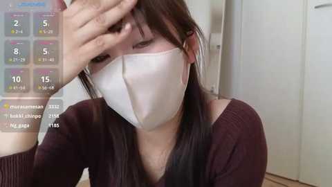 Media: Video of an East Asian woman with long black hair, wearing a white surgical mask and a dark brown sweater. She's sitting, with a hand covering her forehead. A digital clock overlay shows time and weather.