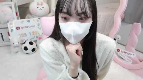 Media: Video of an Asian woman with long black hair, wearing a white mask, pink plush robe, and pink gaming chair in a pastel-themed room with toys and a soccer ball.