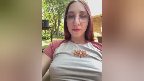 Media: A video of a young woman with fair skin, long red hair, and glasses, wearing a white and pink t-shirt with a sun graphic and the word \"MOM.\" She stands outdoors, with lush green trees and a brick building in the background.