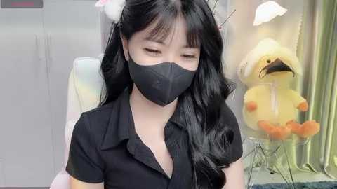 Media: Video of a young Asian woman with long black hair and bangs, wearing a black face mask and black blouse, seated in a room with a stuffed yellow duck and white lamp.