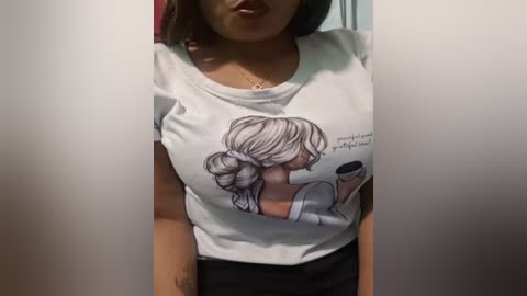 Media: Video of a woman with medium skin tone wearing a white T-shirt featuring a cartoon illustration of a woman with a bun hairstyle holding a cup. She's indoors with a blurred background.