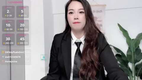 Media: Video of an Asian woman with long, wavy brown hair, wearing a black suit and white shirt, in an office setting with a potted plant and a temperature gauge.