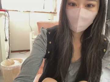 Media: Video of an East Asian woman with long black hair, wearing a gray sweater, black vest, and a pink face mask, sitting in a brightly lit room with beige walls, a white chair, and a basket.