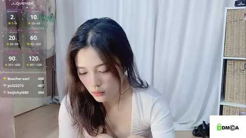 Media: Video of an Asian woman with long black hair, fair skin, and wearing a white top, sitting indoors with a neutral expression.