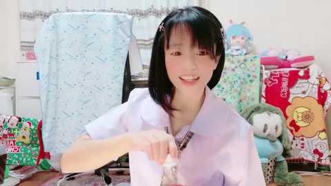 Media: A video of a smiling young Asian woman with straight black hair, wearing a white lab coat, standing in a brightly lit, cluttered room filled with colorful plush toys and boxes.