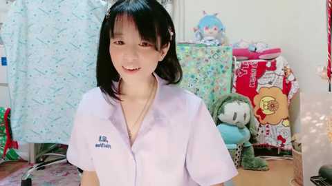 Media: Video of a smiling, light-skinned Asian woman with straight black hair, wearing a white, short-sleeved, collared shirt, in a cluttered, brightly decorated room with plush toys and fabric.