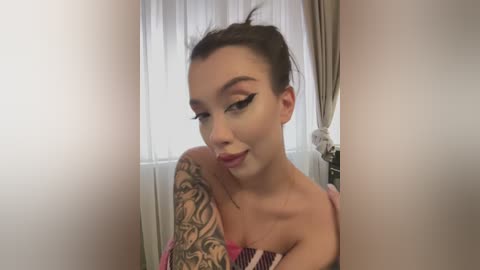 Media: Video of a young woman with light skin, dark hair tied back, wearing a pink top and black eyeliner, standing in a softly lit room with white curtains.