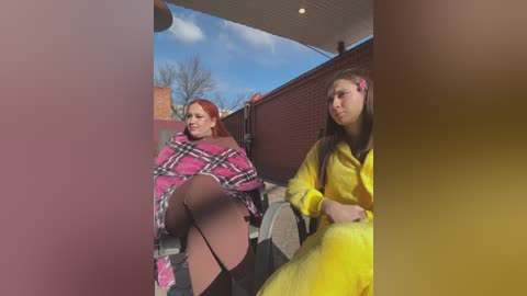 Media: Video of a red-haired woman in a pink plaid blanket, and a brunette in a yellow costume, sitting on a bench outside, with a brick wall and leafless trees in the background.