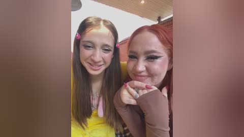 Media: Video of two women with fair skin and long hair, one with red, the other brunette, smiling. Background features a white ceiling and a blurred red curtain.