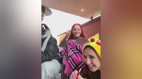 Media: A video showing three people, two women and one man, smiling in a car. The women wear vibrant hats and colorful coats, while the man is in a gray jacket. The background features a brick wall and a bright sky.