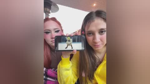 Media: Video of two young women with long hair, one red and one brunette, wearing bright yellow jackets, taking a selfie with a phone displaying a cartoon character.