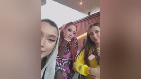 Media: Video of three young women posing outdoors; one with long pink hair, another with long brown hair, and a third with long dark hair, smiling. Red brick building and overcast sky in the background.