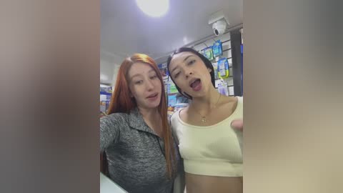 Video of two women inside a brightly lit, modern grocery store. One with long red hair, wearing a gray sweater, smiles. The other with short black hair, wearing a white crop top, poses with tongue out.