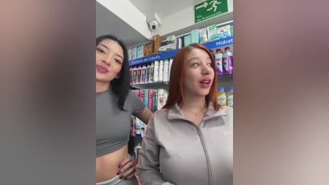 Media: Video of two young women in a pharmacy; one Asian with black hair and red lipstick, wearing a grey crop top, the other Caucasian with red hair, wearing a light grey hoodie. Shelves of products in the background.