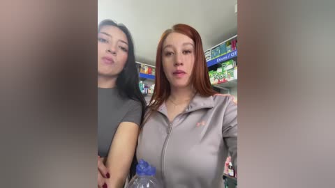 Media: Video of two young women in a store, one with long black hair, the other with red hair, both in grey jackets, holding water bottles, smiling, blurred background.