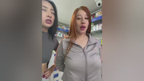 Media: Video of two young women in a pharmacy: one with long black hair in a grey top, the other with red hair in a light jacket. Shelves stocked with medicine in the background.