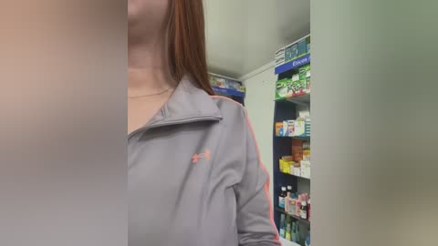 Media: Video of a person with shoulder-length brown hair, wearing a gray Adidas track jacket, standing in a pharmacy with shelves filled with various medications and products.