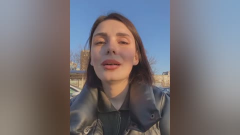 Media: A video of a young woman with fair skin and straight brown hair, wearing a black leather jacket, standing outdoors against a clear blue sky and urban backdrop.