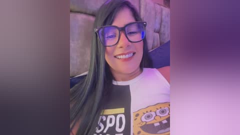 Media: Video of a cheerful, light-skinned woman with long black hair and glasses, wearing a SpongeBob T-shirt, sitting against a cushioned backdrop.