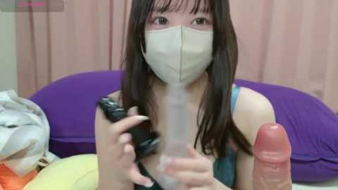 A video of a young Asian woman with long black hair, wearing a surgical mask and a teal tank top, holding a large erect penis in her hands. She is indoors with a purple pillow and a white blanket in the background.