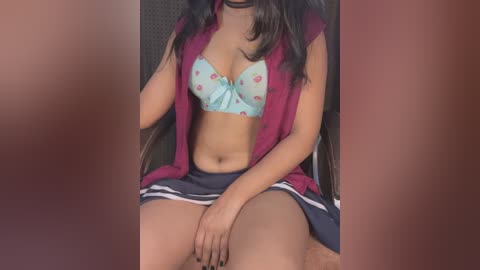 Media: A video of a young woman with medium brown skin, wearing a pink sleeveless shirt, a blue polka dot bra, and a short navy blue skirt with white stripes. She sits on a black chair, her legs spread apart, revealing her thighs.