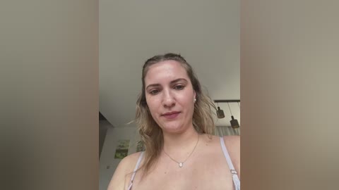 Media: Video of a fair-skinned, blonde woman with light makeup, wearing a white spaghetti strap top, standing indoors with a neutral background.