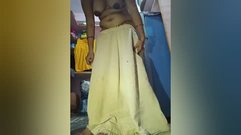 Video of a topless, dark-skinned woman with medium-sized breasts, wearing a yellow sari, standing in a cluttered, dimly lit room with shelves and plastic bags in the background.