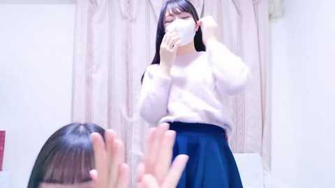 Media: Video of an East Asian woman with long black hair, wearing a white cardigan and blue skirt, blowing her nose in a white tissue, reflected in a mirror, in a white room with beige curtains.