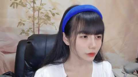 Media: Video of an Asian woman with long black hair, fair skin, and wearing a blue headband, sitting in a black leather chair in a room with beige wall art and a plant.